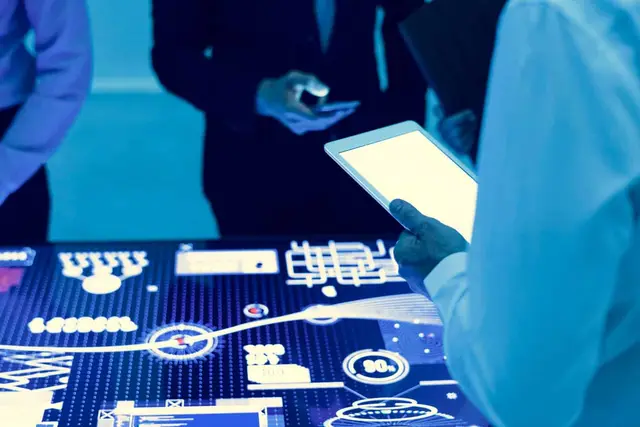 Personas con tableta y teléfono frente a una mesa luminosa con gráficos e indicadores en tonos azules