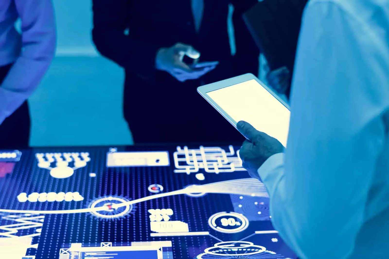 Personas con tableta y teléfono frente a una mesa luminosa con gráficos e indicadores en tonos azules