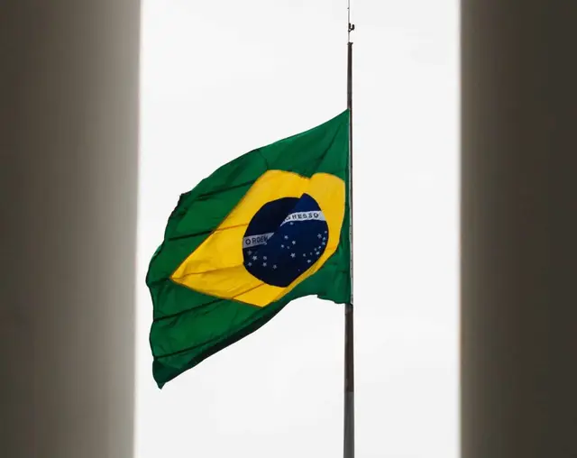 Bandera de Brasil izada a media asta contra un cielo blanco, entre dos muros oscuros