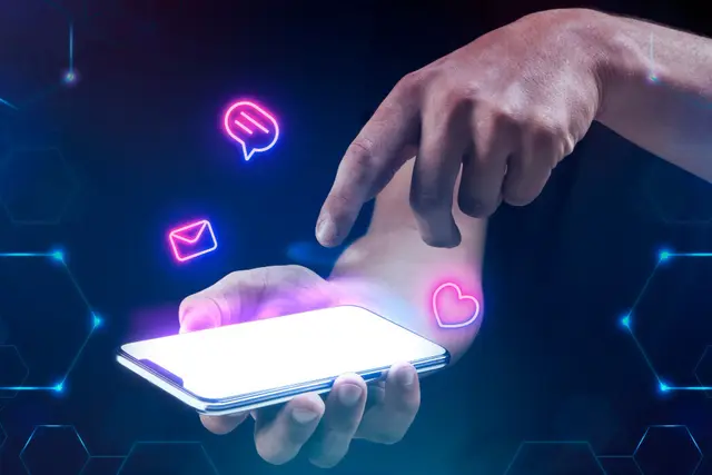 Mano que toca la pantalla de un teléfono con íconos de mensaje, correo y corazón en neón rosa que flotan sobre fondo azul oscuro.
