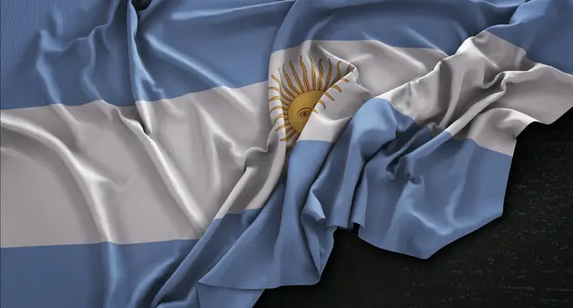 Bandera argentina celeste y blanca con el sol dorado, arrugada y apoyada sobre un fondo oscuro.