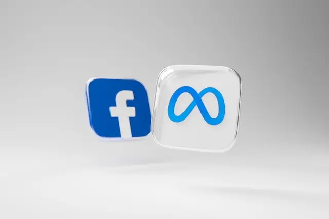Dos íconos 3D flotantes sobre fondo gris claro: el logotipo de Facebook en azul y el símbolo infinito de Meta en un cubo translúcido.