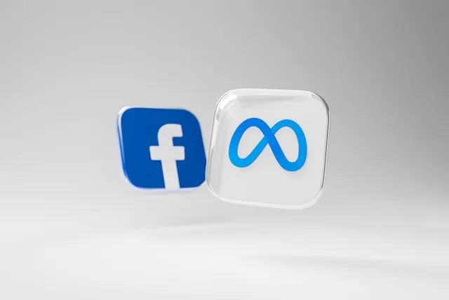 Dos íconos 3D flotantes sobre fondo gris claro: el logotipo de Facebook en azul y el símbolo infinito de Meta en un cubo translúcido.