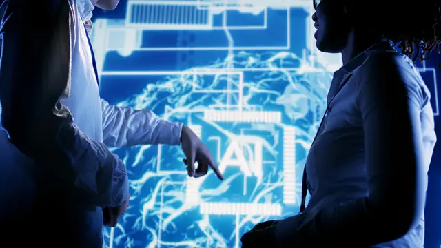 Dos personas de pie frente a una pantalla azul con las letras AI sobre un cerebro luminoso, una de ellas señala el centro con la mano.