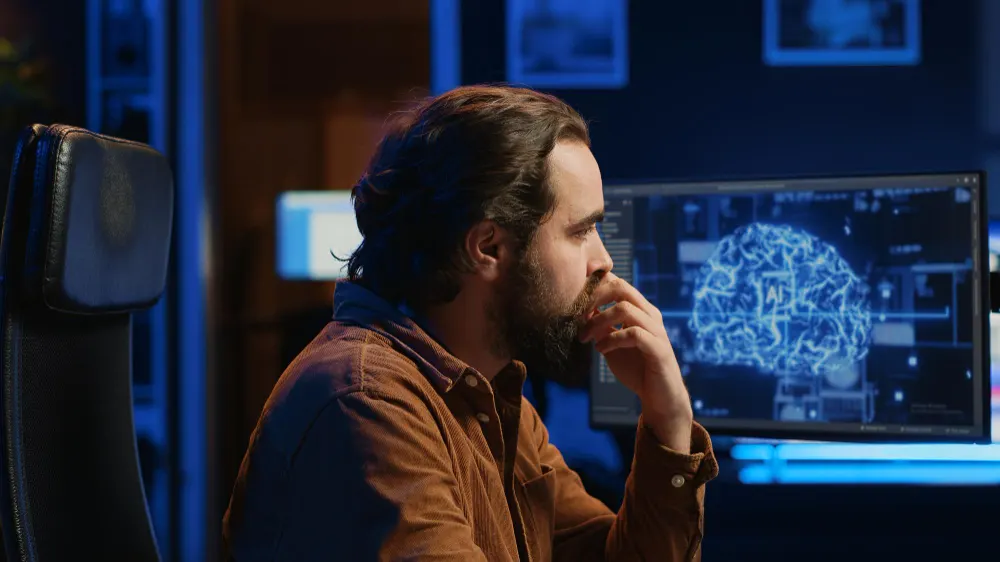 Hombre barbudo con la mano en la boca mira un monitor con la imagen de un cerebro azul, en una oficina de luz tenue.