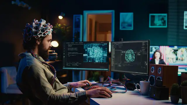 Hombre con un casco de electrodos frente a dos monitores que muestran imágenes de un cerebro, en una oficina en penumbra.