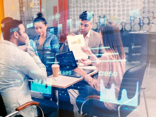Cuatro profesionales trabajan con laptops y papeles en una mesa, vistos a través de un vidrio sobre el que flotan gráficos y cifras luminosas.