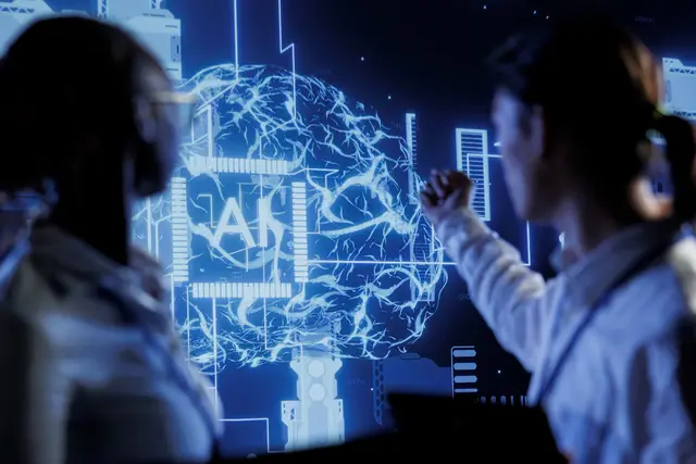 Dos personas frente a una pantalla azul que muestra un cerebro de líneas luminosas con las letras AI en el centro.