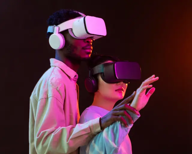 Dos personas con visores de realidad virtual y auriculares bajo luz rosada y azul; una levanta la mano como si tocara algo frente a ella.