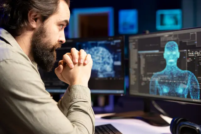 Hombre barbudo de perfil con las manos entrelazadas ante dos monitores con un cerebro digital y una figura humana en 3D