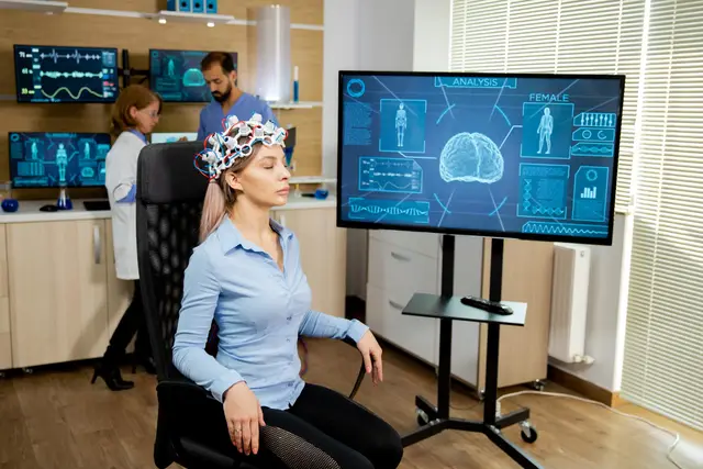 Mujer con un gorro de electrodos que registra su actividad cerebral, frente a una pantalla con un cerebro y datos; dos investigadores trabajan al fondo.