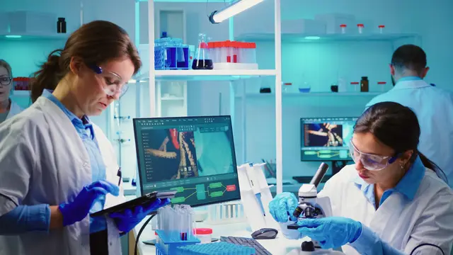 Dos científicas con bata y guantes trabajan en un laboratorio iluminado de azul, con una tableta, un microscopio y un monitor