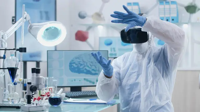 Persona con traje de protección y casco de realidad virtual levanta las manos con guantes ante un monitor que muestra un cerebro
