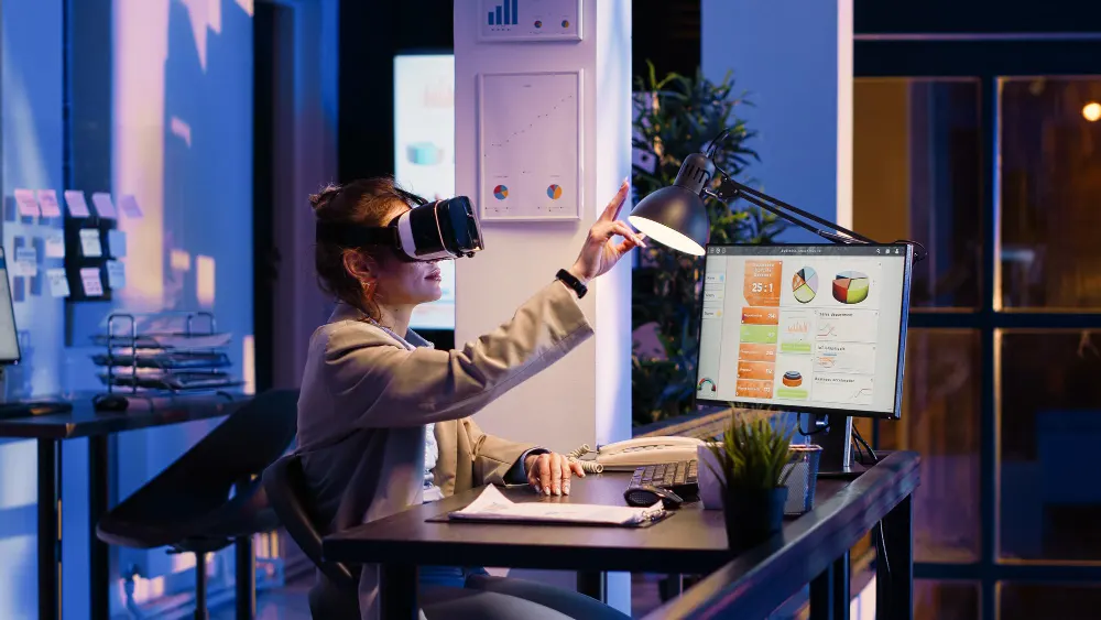 Mujer con visor de realidad virtual que levanta la mano ante un escritorio con un monitor de gráficos, en una oficina de noche