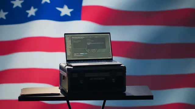 Laptop abierta sobre un maletín negro en una mesa pequeña, frente a una gran bandera de Estados Unidos.