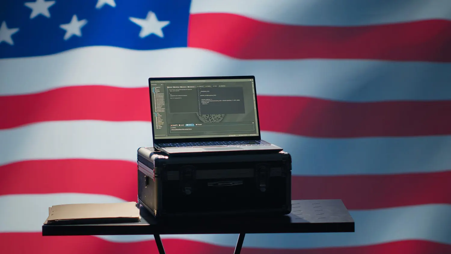 Laptop abierta sobre un maletín negro en una mesa pequeña, frente a una gran bandera de Estados Unidos.