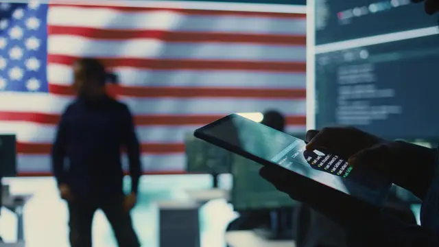 Silueta de un hombre frente a una bandera de Estados Unidos y pantallas con código, con una tableta en primer plano
