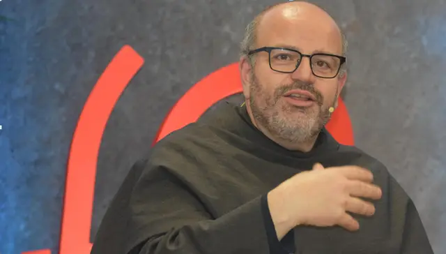Primer plano de un fraile franciscano con gafas y hábito oscuro que habla y gesticula ante un fondo con formas rojas