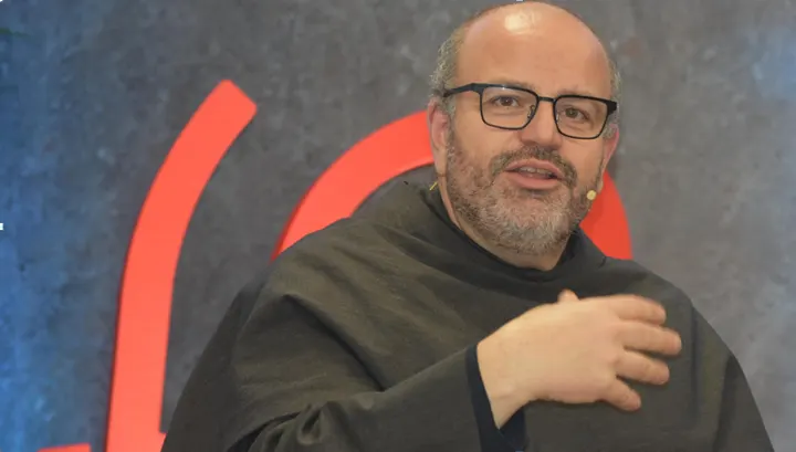 Primer plano de un fraile franciscano con gafas y hábito oscuro que habla y gesticula ante un fondo con formas rojas