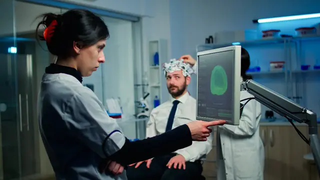 Una científica señala con el dedo una pantalla con la imagen de un cerebro verde mientras un colega con casco de electrodos espera sentado
