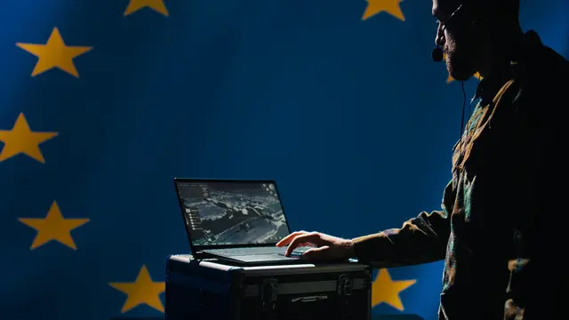Hombre con auriculares escribe en una laptop frente a una bandera azul de la Unión Europea con estrellas amarillas