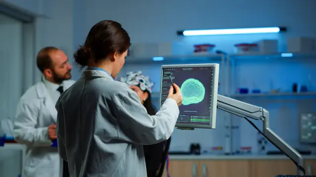 Una científica señala una pantalla con la imagen de un cerebro verde mientras un colega con casco de electrodos la observa en un laboratorio