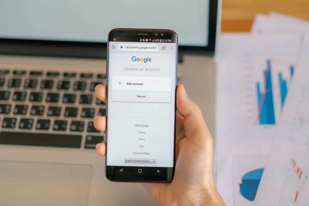 Mano que sostiene un teléfono con la pantalla de acceso de Google, junto a un portátil y gráficos impresos