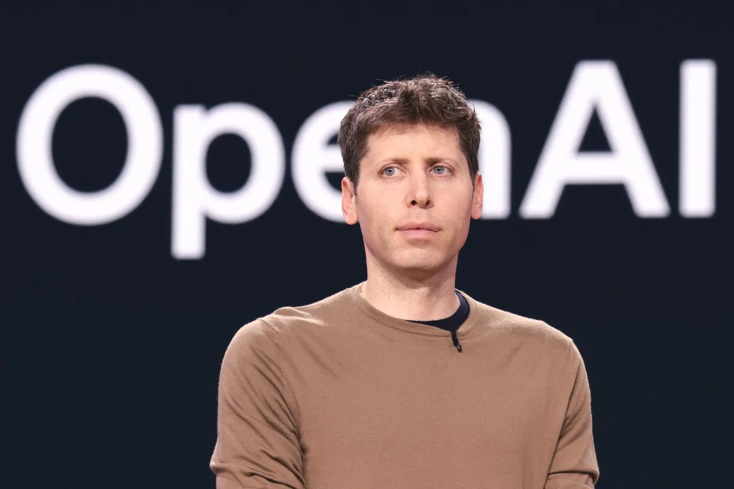 Sam Altman con camiseta marrón frente a un fondo oscuro con el logotipo de OpenAI