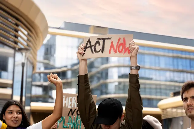 Manifestantes con un cartel que dice «Act Now» frente a edificios de oficinas modernos
