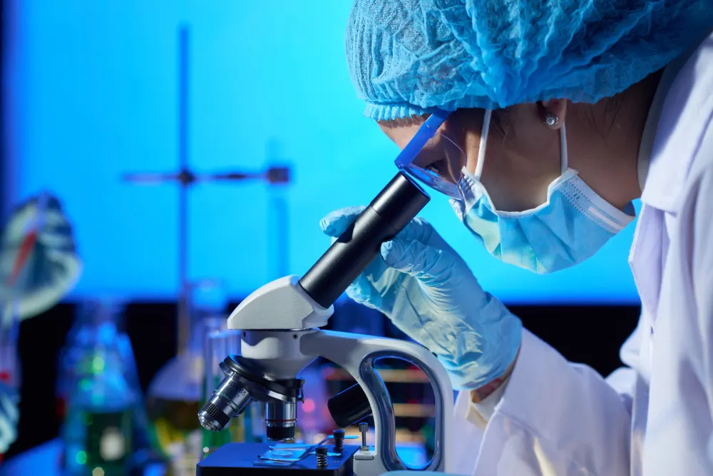 Investigadora con gorro azul y mascarilla mira por un microscopio en un laboratorio iluminado de azul