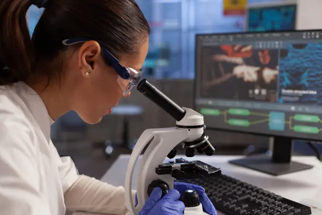 Investigadora con gafas de protección mira por un microscopio, junto a un monitor con imágenes de células, en un laboratorio