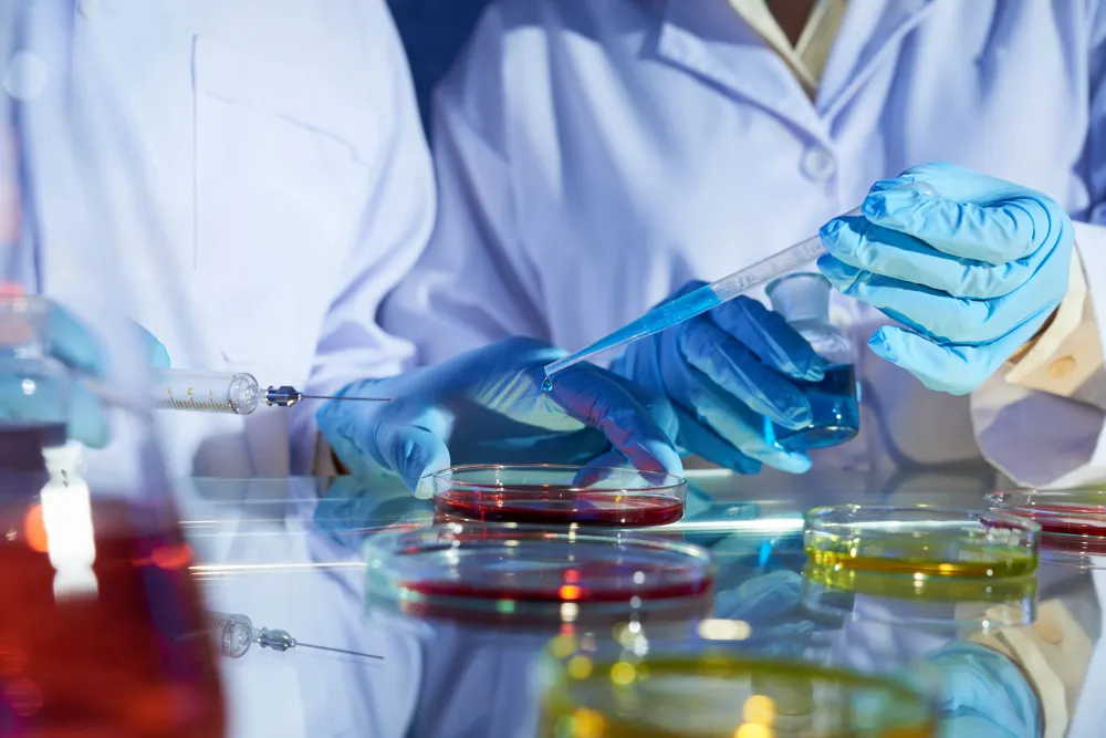 Manos con guantes azules y bata blanca manipulan una pipeta sobre placas de Petri con líquidos rojos y amarillos en un laboratorio