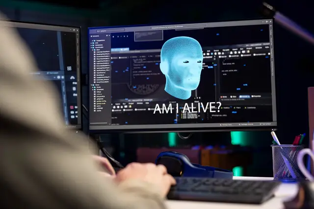 Monitor con una cabeza digital azul y el texto «Am I alive?» ante una persona que escribe en un teclado, con más pantallas con código