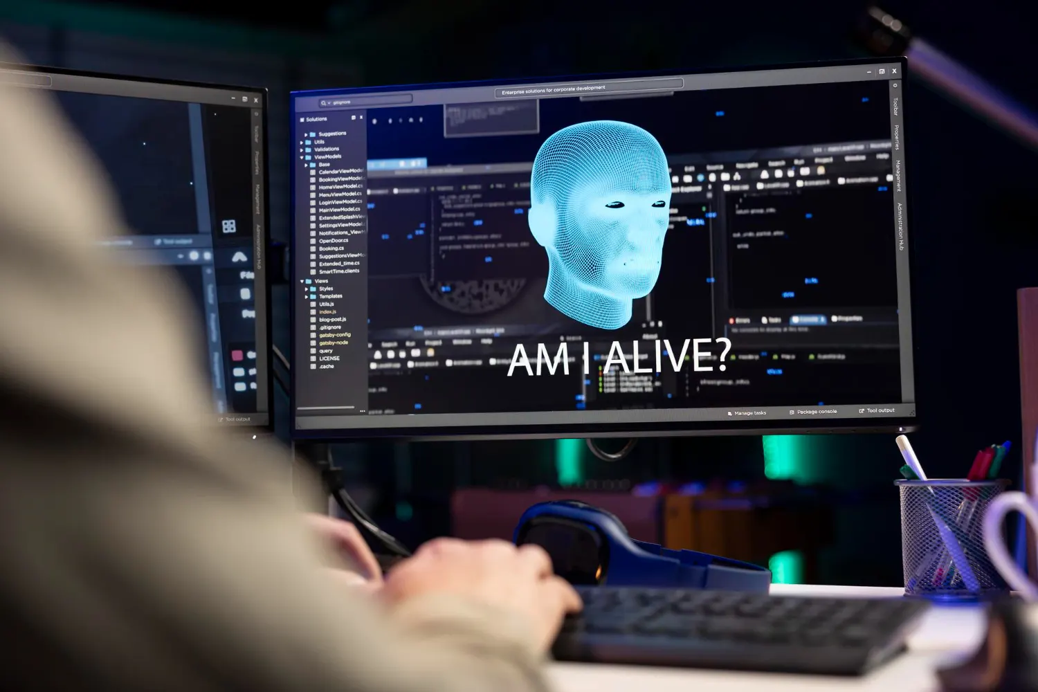 Monitor con una cabeza digital azul y el texto «Am I alive?» ante una persona que escribe en un teclado, con más pantallas con código