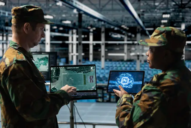 Dos militares con uniforme de camuflaje consultan mapas y una pantalla con un cerebro digital en sus portátiles, dentro de un gran hangar