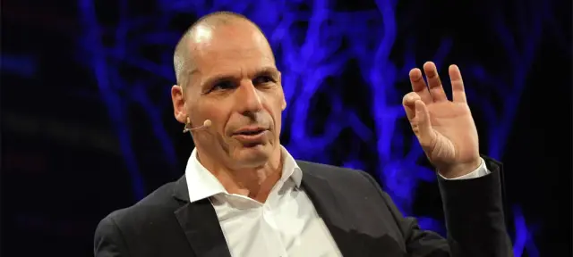 Yanis Varoufakis habla sobre un escenario, con micrófono de diadema y una mano en alto, frente a un fondo azul oscuro con luces