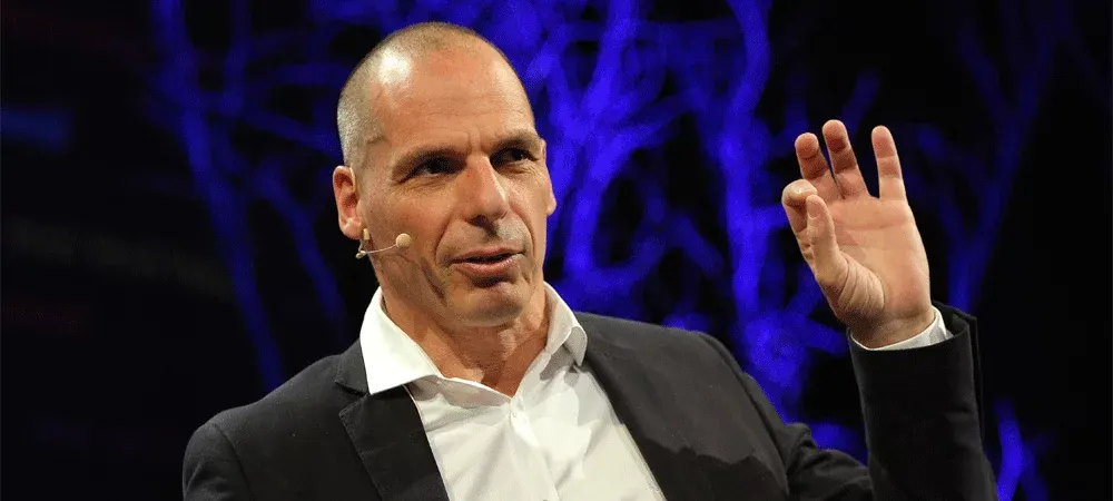 Yanis Varoufakis habla sobre un escenario, con micrófono de diadema y una mano en alto, frente a un fondo azul oscuro con luces