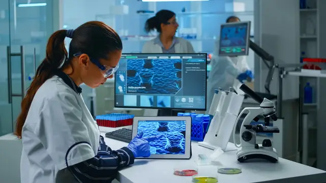 Científica con bata, guantes y gafas de seguridad que observa una tableta y un monitor con imágenes microscópicas azules en un laboratorio