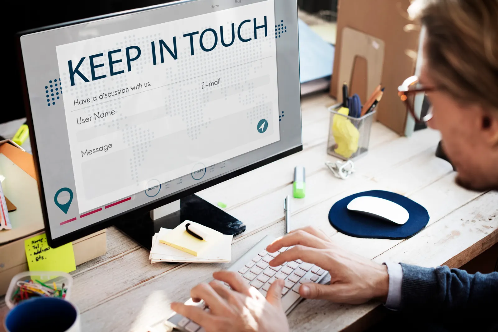 Hombre con gafas que escribe en un teclado frente a un monitor con un formulario de contacto titulado «Keep in touch»