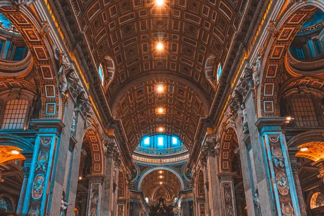 Interior de una basílica con bóveda artesonada, arcos y columnas ornamentadas bajo luces cálidas