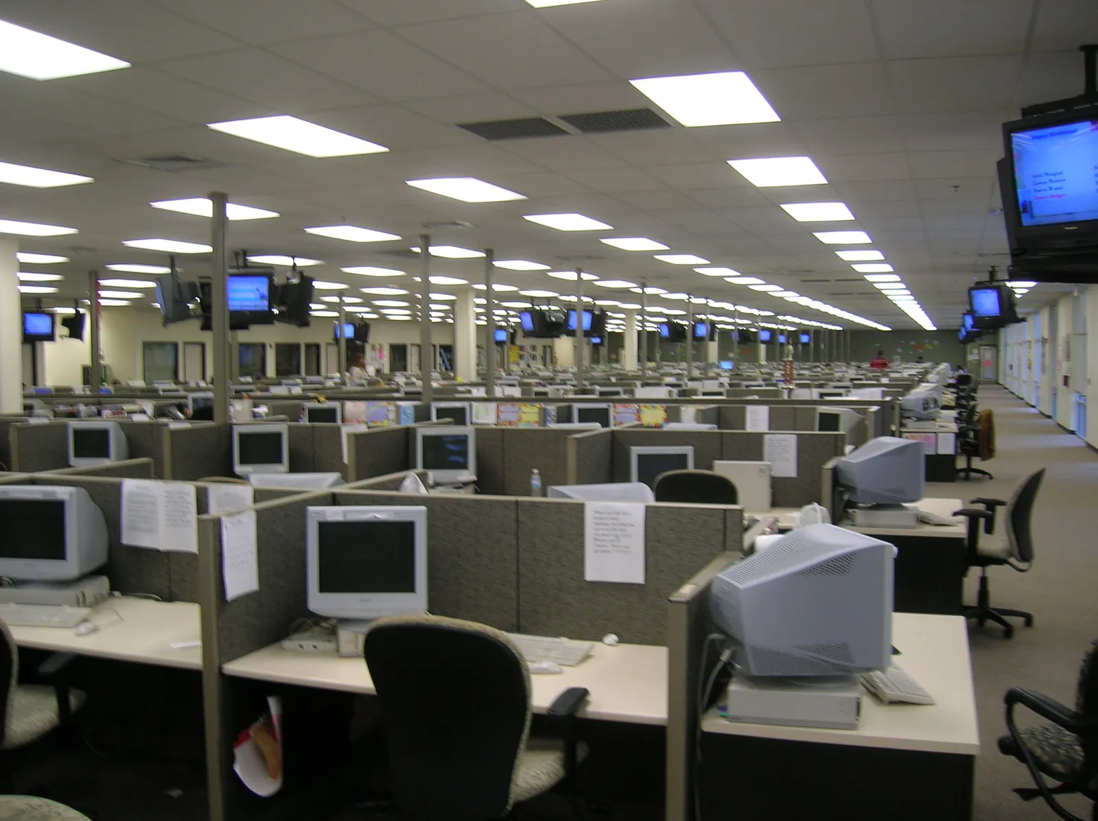 Filas de cubículos con monitores de tubo, teclados y sillas en una amplia oficina con luces rectangulares de techo.